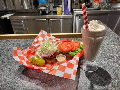 A burger with lettuce, tomato, sprouts, pickles, and a milkshake with whipped cream on a checkered tray.
