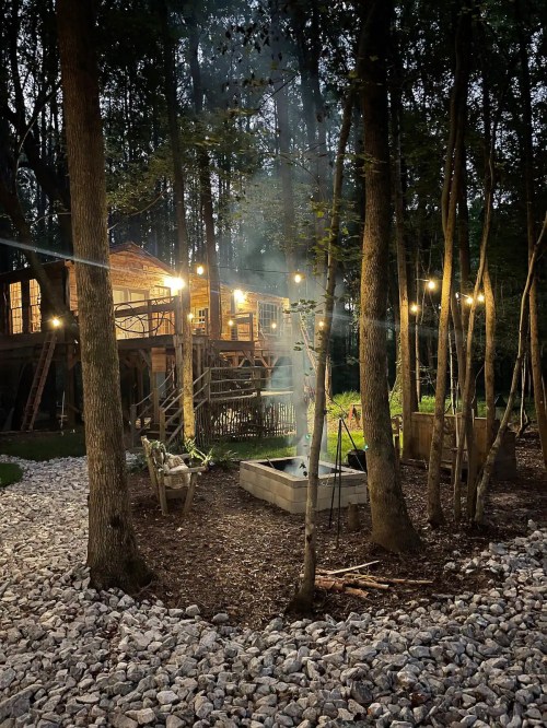 A cozy cabin in the woods at night, surrounded by trees and illuminated by string lights, with a fire pit in the foreground.