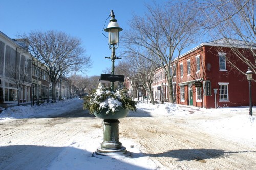 A snowy street scene featuring a decorative lamp post and quaint buildings under a clear blue sky.