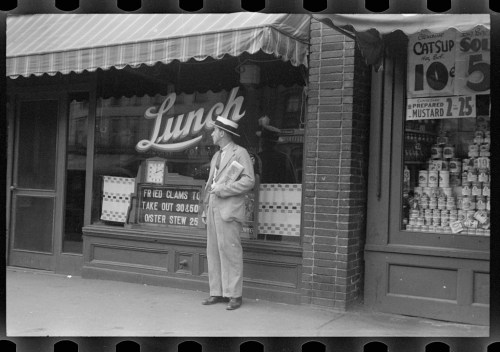 A man in a suit and hat stands outside a lunch counter with a sign displaying menu items and prices.