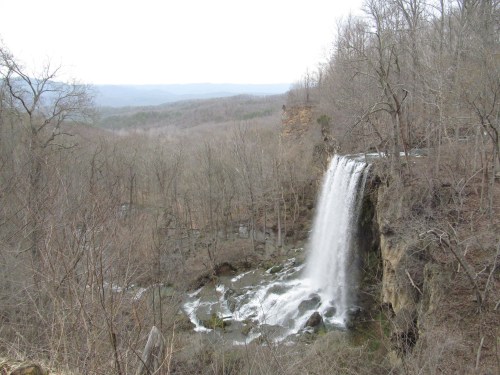 A waterfall cascades over rocky cliffs, surrounded by bare trees and distant mountains under a cloudy sky.