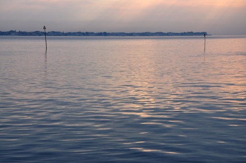 Calm water reflects soft light, with distant land and two poles marking the shoreline under a pastel sky.