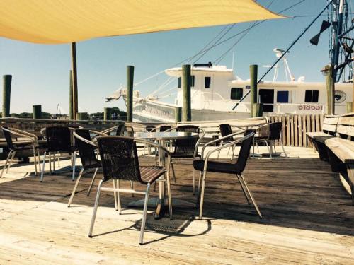 Outdoor seating area with empty tables and chairs, overlooking a boat docked in the background under a sunny sky.
