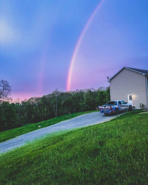 A truck parked on a gravel driveway beside a house, with a vibrant rainbow arching over a green landscape at sunset.