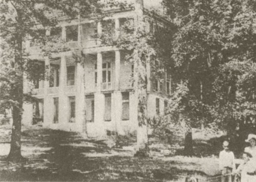 A historic, two-story house with columns, surrounded by trees, with a few people visible in the foreground.