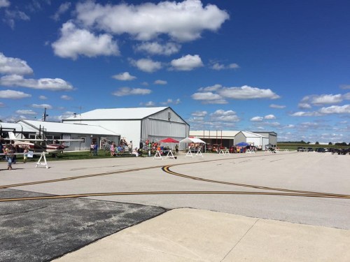 A sunny day at an airfield with hangars, people, and tents set up along the runway under a blue sky with clouds.