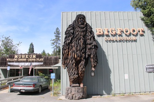A wooden Bigfoot statue stands outside a museum, with a sign reading "Bigfoot Collection" and a vintage car nearby.