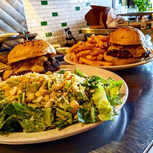 Two burgers with cheese and sesame seed buns, served with fries and a side salad on a metal table.