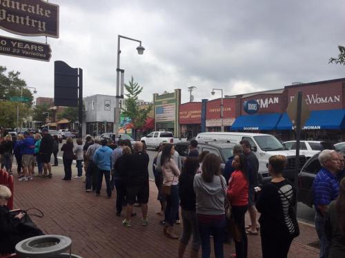 A long line of people waiting outside shops on a cloudy day in a busy street.