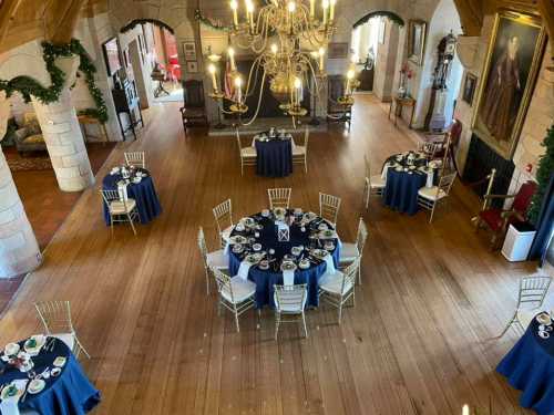 Elegant dining setup in a spacious hall, featuring round tables with blue tablecloths and fine china, surrounded by festive decor.