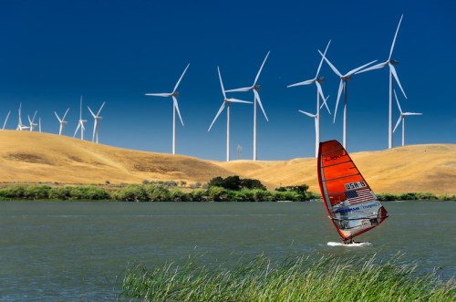 A windsurfer glides across a lake with wind turbines and rolling hills in the background under a clear blue sky.