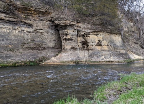 A serene river flows beside a rocky cliff, surrounded by greenery and trees in a peaceful natural setting.