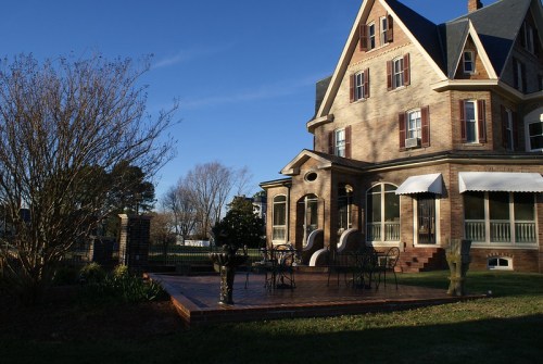 A large, historic house with a wraparound porch and a wooden deck, surrounded by trees and a clear blue sky.