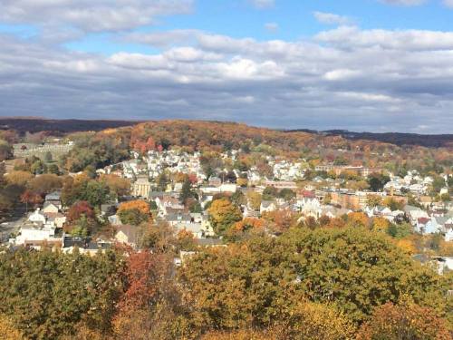 A scenic view of a town surrounded by vibrant autumn foliage and rolling hills under a partly cloudy sky.