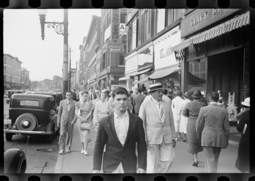 A busy street scene from the past, featuring pedestrians and vintage cars, with shops lining the sidewalk.