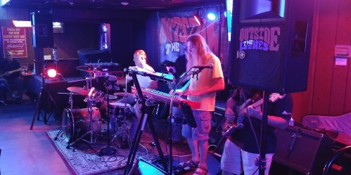 A live band performs on stage with drums, keyboards, and guitars in a dimly lit venue. Colorful lights illuminate the scene.