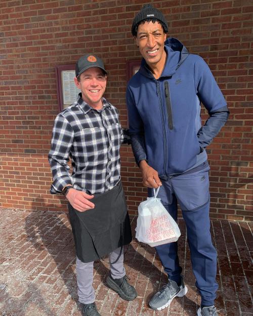 Two men stand outside a building, one in a checkered shirt and apron, the other in a blue tracksuit holding a bag.