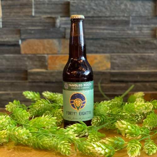 A bottle of Tangled Roots Brewing Company's Forty? Eighty? beer, surrounded by green foliage on a wooden surface.