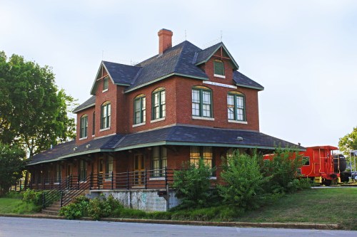 Historic brick building with large windows, a steep roof, and a red train car nearby, surrounded by greenery.