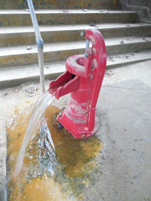 A red hand pump with water flowing from it, set on a concrete surface near stone steps.