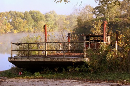 Abandoned concrete platform by a misty river, surrounded by overgrown vegetation and autumn foliage.