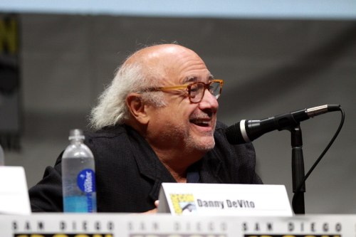 A smiling man with glasses speaks into a microphone at a panel, with a water bottle in front of him.