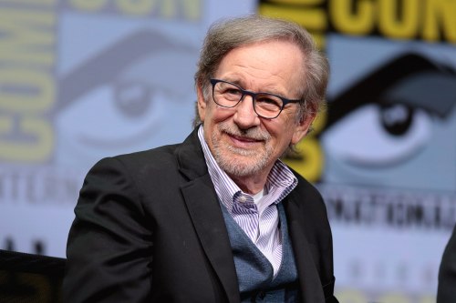 A smiling man with glasses and gray hair sits on stage, wearing a black suit and blue sweater, at a convention event.