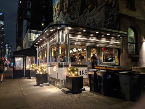 A brightly lit diner at night, featuring a modern design and outdoor seating, with city lights in the background.