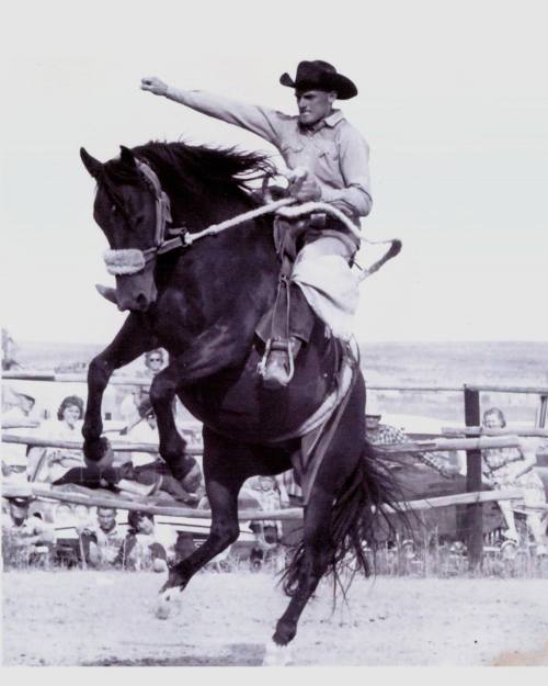 A cowboy riding a bucking horse, raising his arm in excitement, with spectators watching in the background.