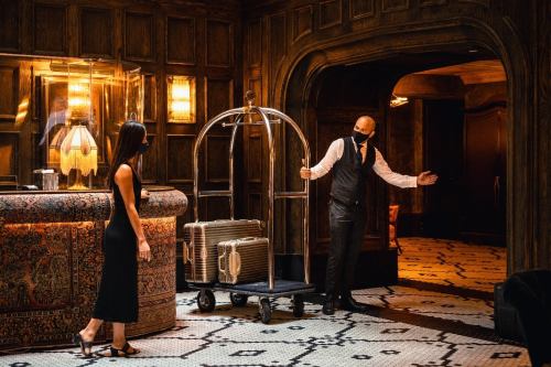 A hotel staff member in a mask greets a guest near a luggage cart in a stylish, wood-paneled lobby.