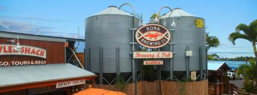 Kona Brewing Co. sign with silver brewing tanks and a tropical setting, featuring palm trees and a clear sky.