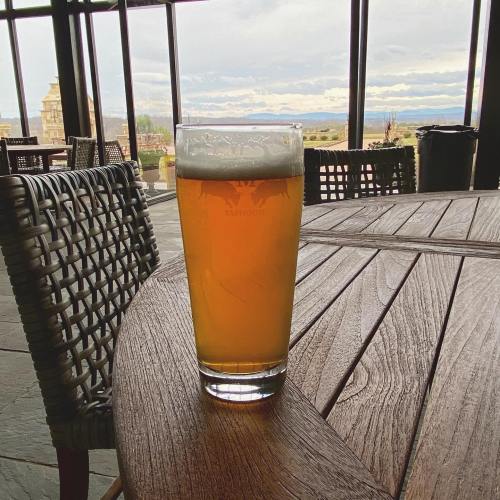 A glass of beer sits on a wooden table with a scenic view of mountains and cloudy skies in the background.