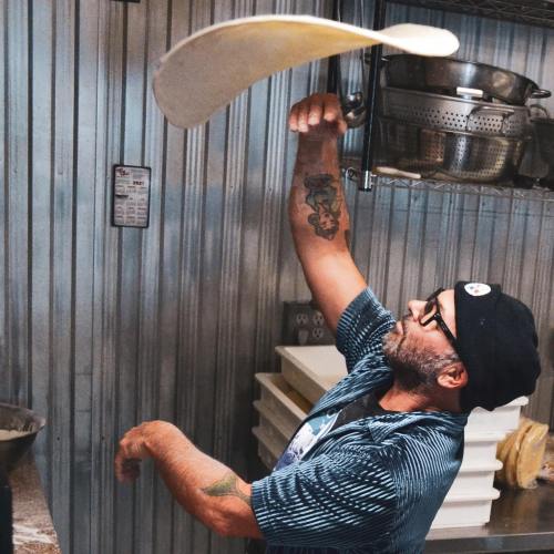 A chef tossing pizza dough in a kitchen, wearing a striped shirt and glasses, with shelves of kitchenware in the background.