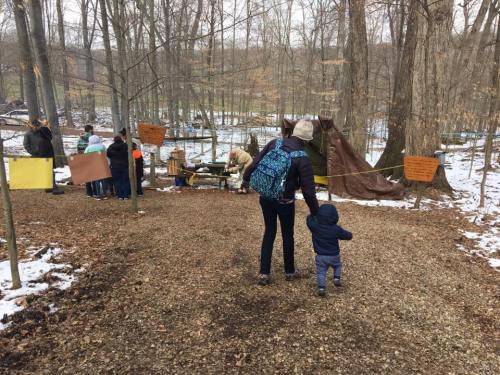 A child and adult walk on a gravel path in a snowy forest, with people and signs in the background.