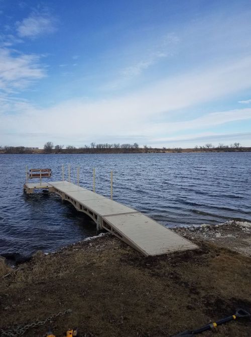 A wooden dock extends into a calm lake under a blue sky with scattered clouds. Grass lines the shore.