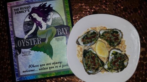 A plate of baked oysters with lemon, next to a colorful menu for Oyster Bay restaurant featuring a mermaid.