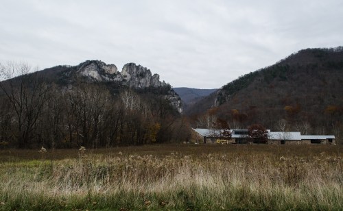 A modern building sits in a grassy field, framed by rocky mountains and overcast skies in a rural landscape.