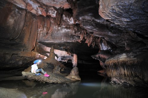 A person sits on a rock in a cave, surrounded by stalactites and a calm water pool, illuminated by soft lighting.