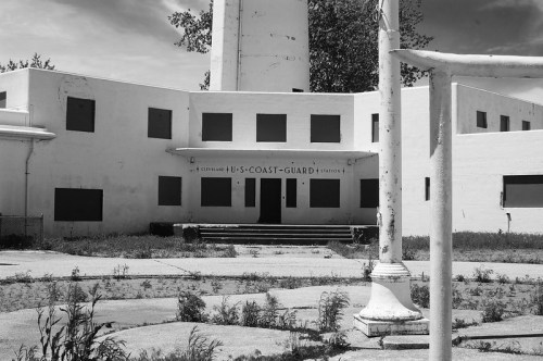 Abandoned U.S. Coast Guard station with boarded windows and overgrown vegetation in black and white.