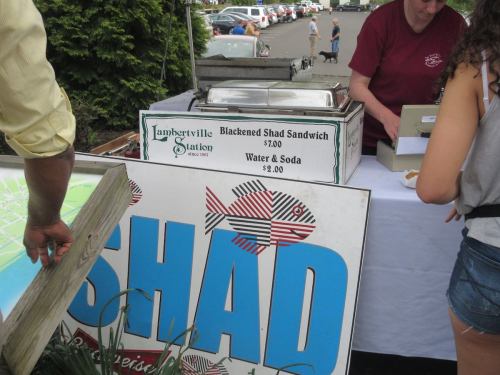 A food stall displaying a menu for blackened shad sandwiches and drinks, with a person taking an order.
