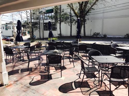 Outdoor café patio with empty black tables and chairs, shaded by umbrellas, surrounded by trees and a quiet street.