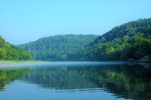 A serene lake surrounded by lush green hills, with morning mist hovering over the water's surface.