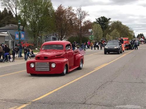 A red vintage truck with an American flag drives down a street lined with spectators during a parade.