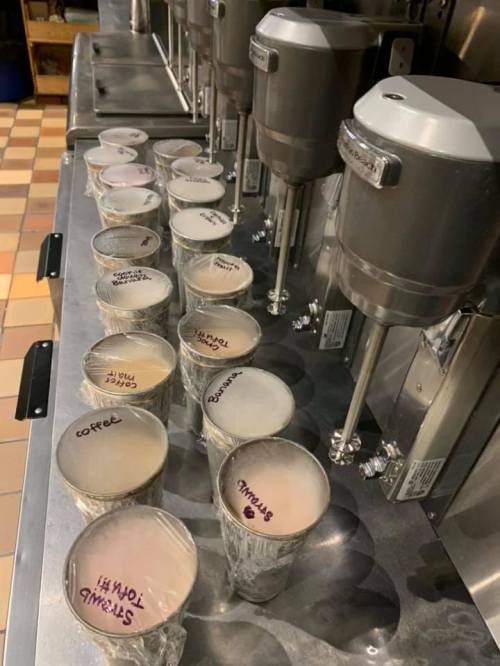 A row of labeled cups filled with different beverages, lined up next to beverage dispensers in a kitchen.