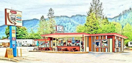 A colorful roadside diner with picnic tables, surrounded by trees and mountains under a cloudy sky.