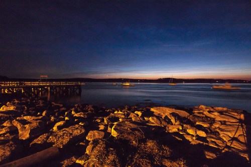 A serene twilight scene by the water, featuring a rocky shore and a pier under a deep blue sky.