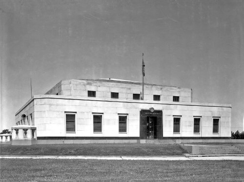 A large, modernist building with a flat roof, featuring large windows and a flagpole in front, set against a clear sky.