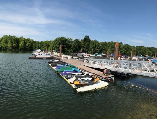 A marina with various boats and jet skis docked at a wooden pier, surrounded by green trees and blue sky.