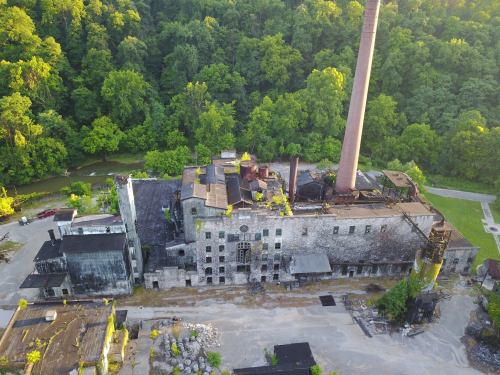 Aerial view of an abandoned industrial building surrounded by lush greenery and a winding creek.