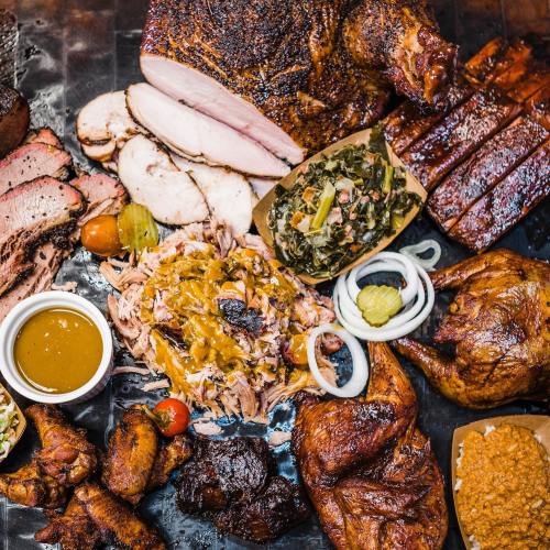 A variety of smoked meats including pulled pork, ribs, chicken, and sides like collard greens and sauce on a platter.